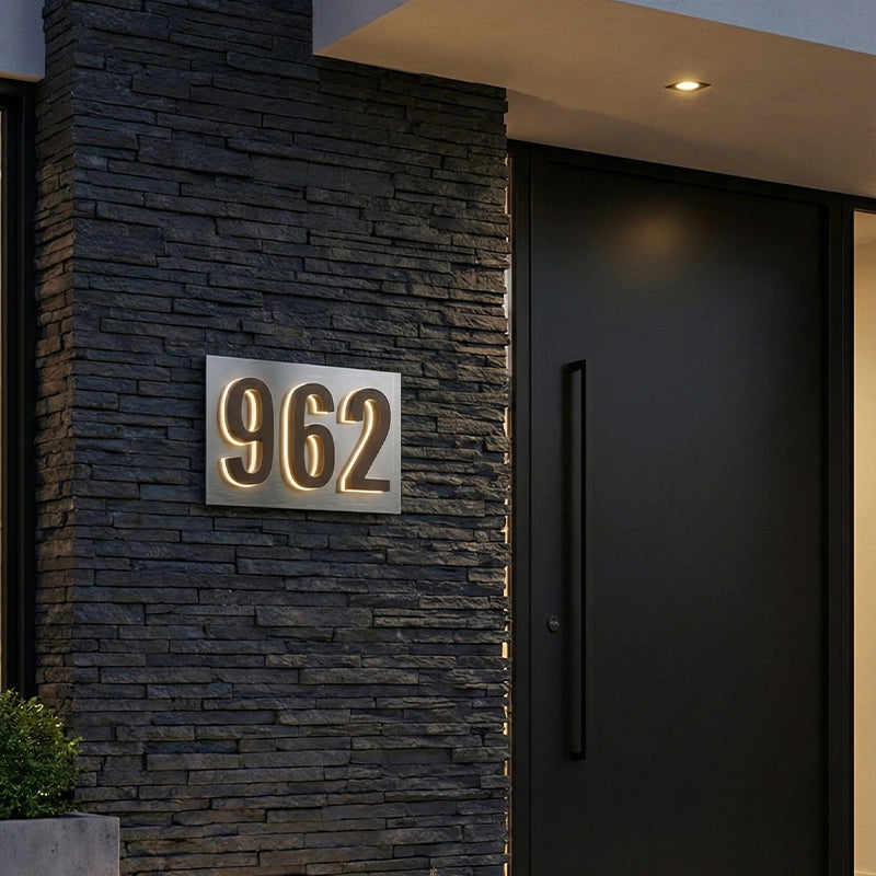 Illuminated house numbers on a brushed mounting plate at dusk